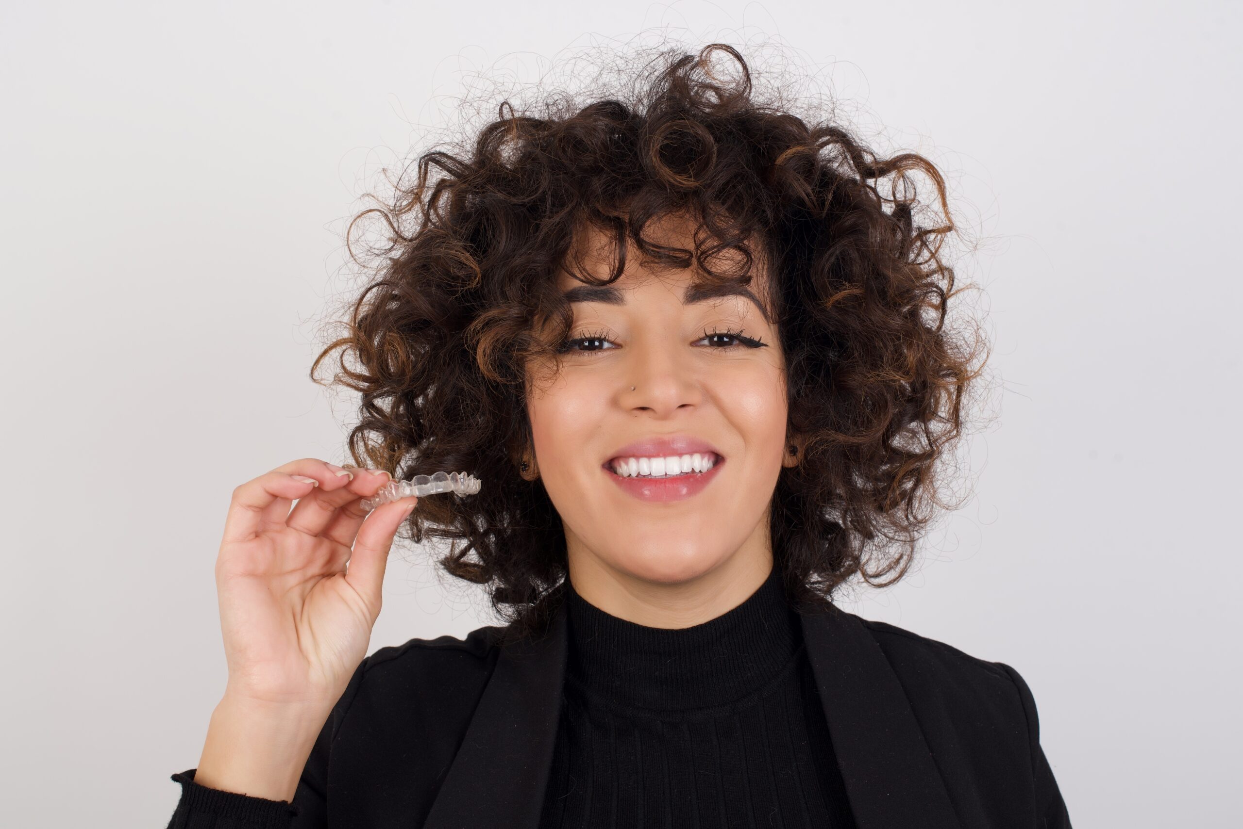 Young beautiful Arab smiling woman with curly hair wearing black suit holding and recommending an invisible retainer for her teeth. Dental healthcare concept.