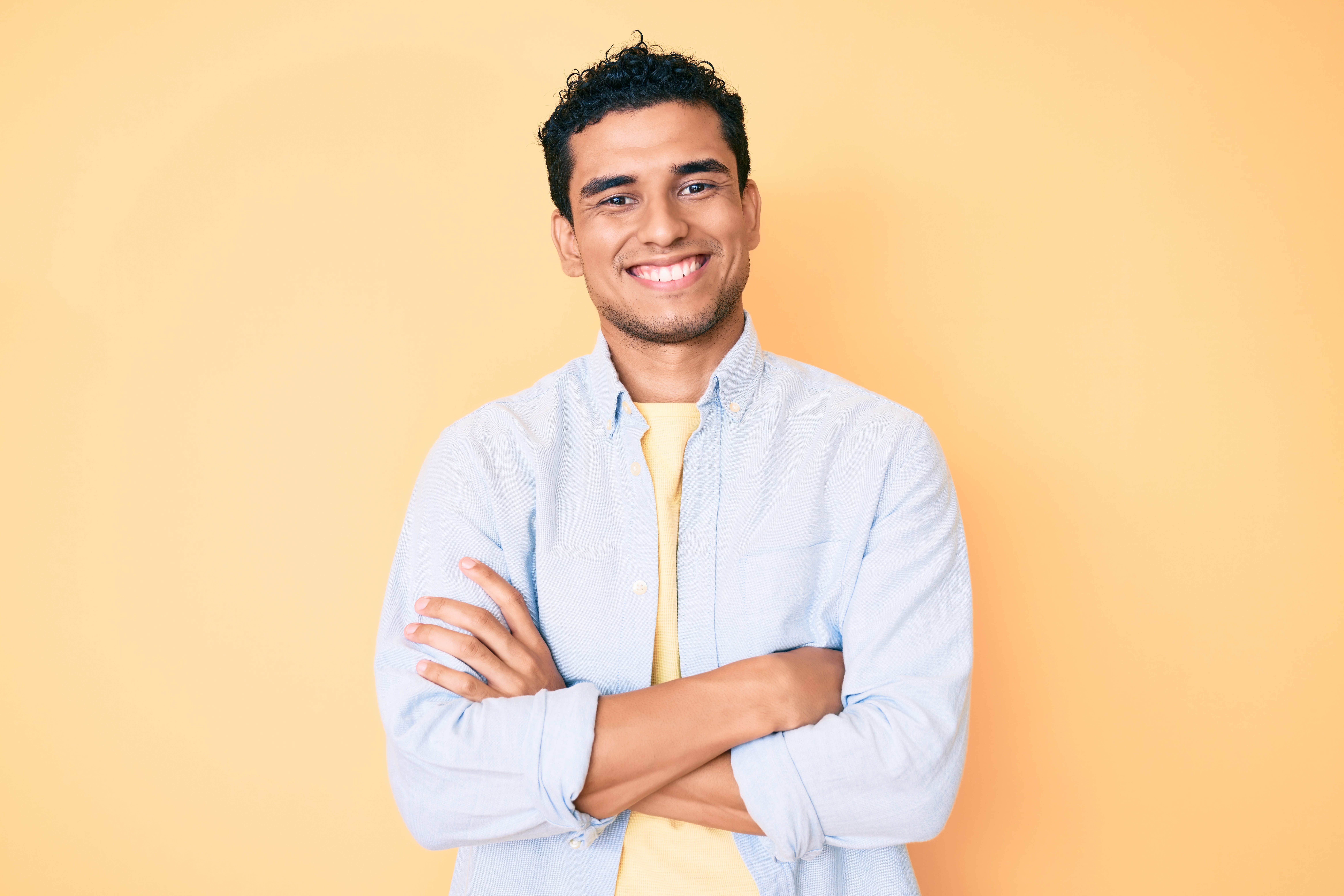 Young handsome hispanic man standing over yellow background happy face smiling with crossed arms looking at the camera. positive person.